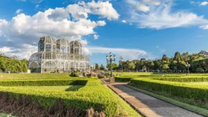 O Jardim Botânico de Curitiba se estende sob um céu azul pontilhado de nuvens brancas e fofas. No centro, a estufa principal, com sua estrutura metálica e vidro, brilha sob o sol. Seus três domos curvos, que lembram palácios de cristal, abrigam uma variedade de plantas. Em primeiro plano, um caminho de pedra ladeado por arbustos bem cuidados convida a um passeio tranquilo. Ao fundo, árvores frondosas completam a paisagem, oferecendo sombra e frescor aos visitantes.