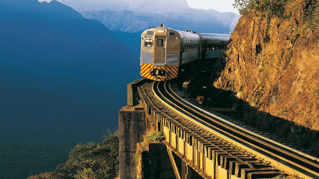 Um trem de passageiros em uma linha férrea que atravessa uma ponte estreita sobre um vale profundo. As montanhas ao fundo criam um cenário dramático.