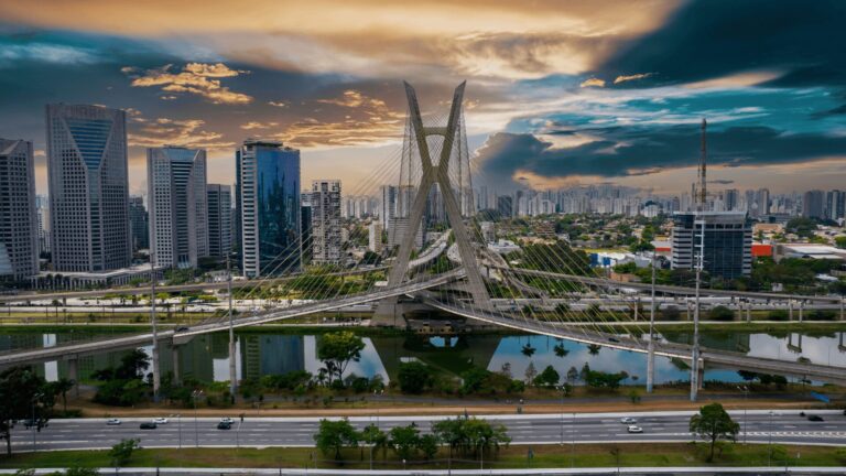 Uma vista aérea espetacular da Ponte Estaiada em São Paulo, Brasil, ao entardecer. A ponte, com seu design moderno e cabos que se estendem como asas, cruza o Rio Pinheiros e se destaca no horizonte urbano da cidade. Prédios altos e modernos se erguem ao fundo, enquanto o céu exibe tons vibrantes de laranja, rosa e roxo, criando um contraste impressionante com a silhueta da ponte e dos edifícios. A água do rio reflete as cores do céu e as luzes da cidade, adicionando um toque mágico à cena.