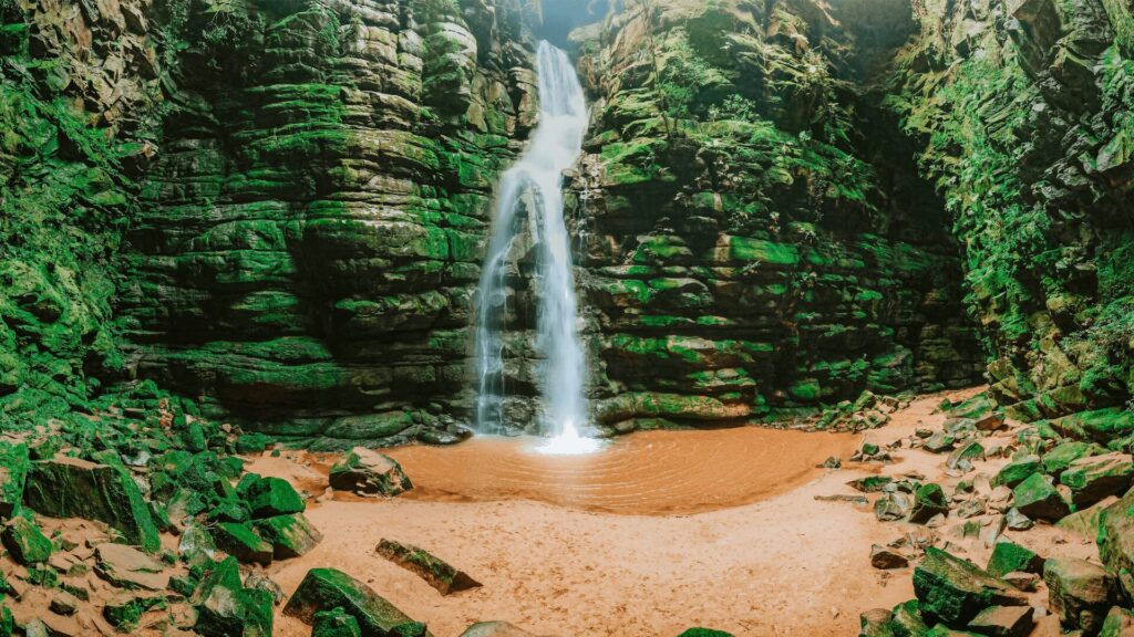 Uma cachoeira majestosa despenca de um paredão rochoso coberto por vegetação verdejante. A água cristalina cai em uma piscina natural de areia clara, formando uma névoa refrescante. As rochas ao redor, também cobertas de musgo, completam a beleza selvagem e intocada do local. A luz do sol que entra pela abertura superior ilumina a cachoeira, criando um contraste de luz e sombra que realça a atmosfera mágica da cena.