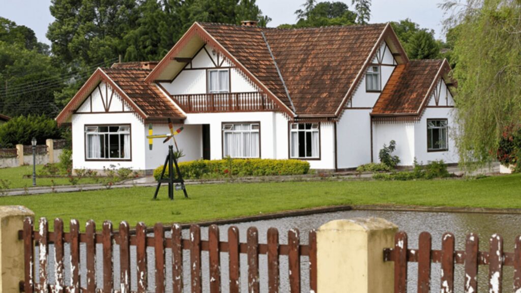 Uma charmosa casa em estilo enxaimel se destaca em um jardim bem cuidado. A fachada branca contrasta com o telhado de telhas marrons e as janelas com molduras escuras. Uma varanda com detalhes em madeira adorna o segundo andar, adicionando um toque rústico. No jardim, um cata-vento colorido gira ao sabor do vento, enquanto um pequeno lago complementa a paisagem serena. A cerca de madeira delimita a propriedade, e ao fundo, árvores altas completam o cenário tranquilo.