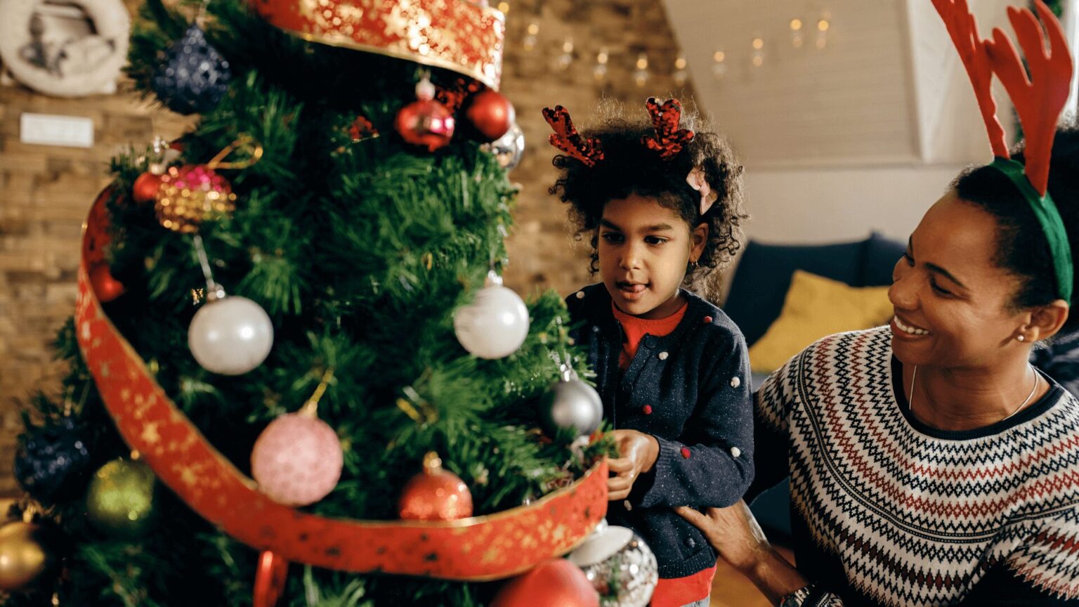 Árvore de Natal decorada com fita vermelha, e duas pessoas em trajes festivos próximas.