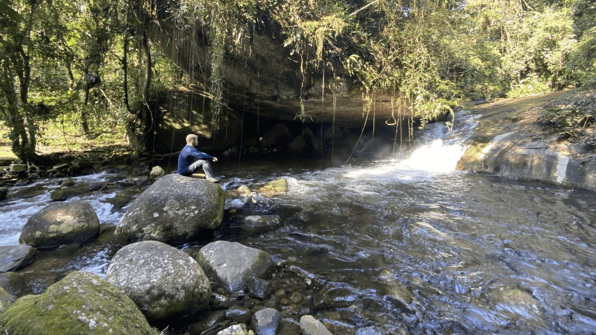 Uma pessoa senta-se em uma pedra perto de uma cachoeira em uma floresta exuberante.