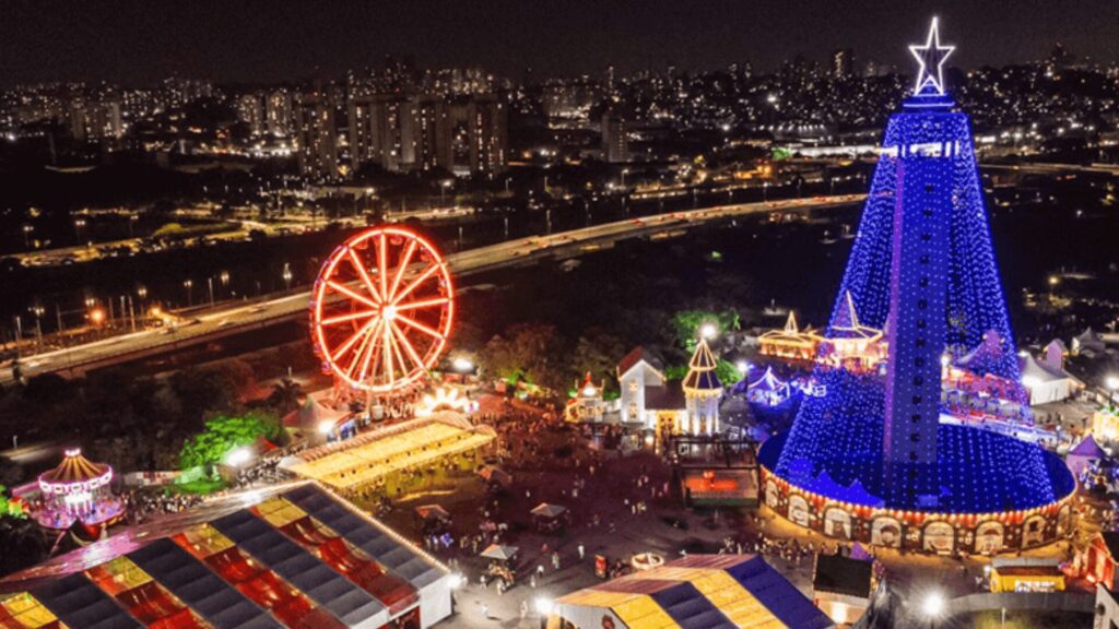 Visão noturna de uma feira festiva com uma roda gigante iluminada e uma grande árvore de Natal iluminada.