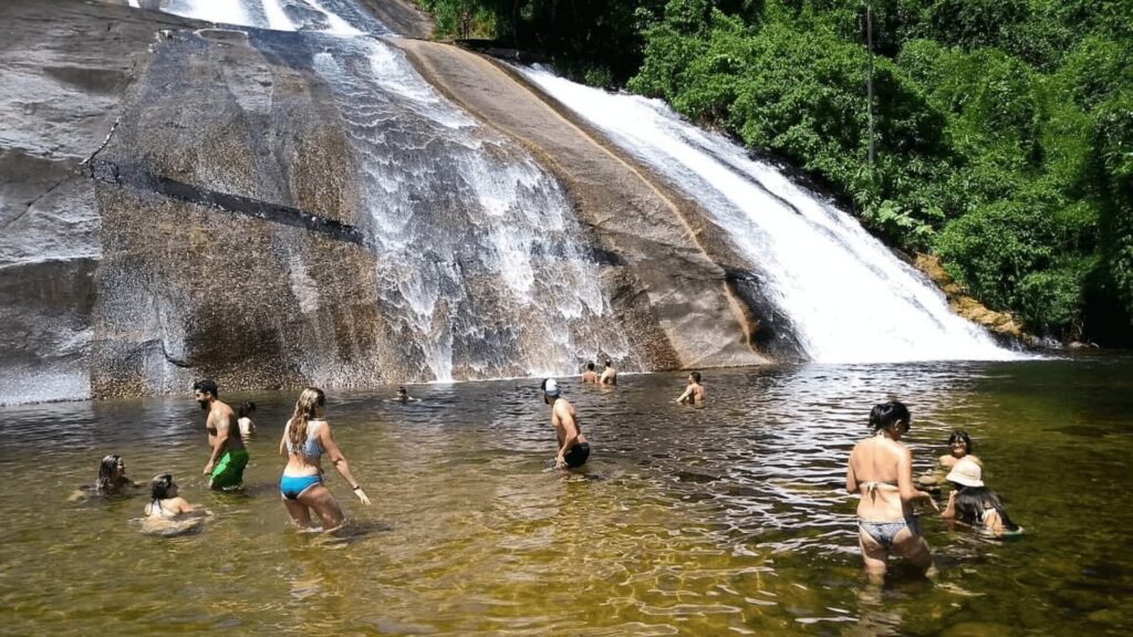 Pessoas nadando na base de uma cachoeira ensolarada cercada por uma vegetação exuberante.