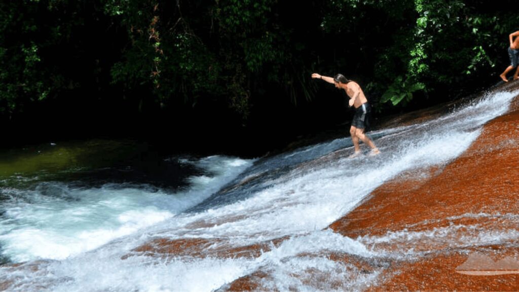 Dois indivíduos deslizando em um toboágua natural em um ambiente florestal.