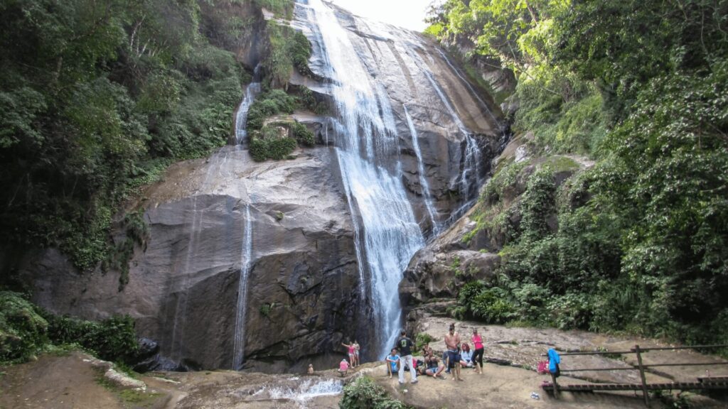 Uma cascata cênica com visitantes na base, cercada por uma vegetação exuberante.