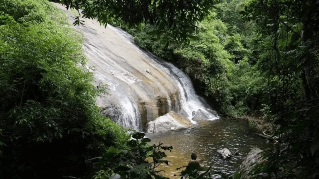 Uma cachoeira serena cercada por uma vegetação exuberante com uma pessoa observando de baixo.