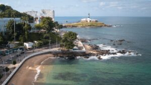 Vista aérea de um litoral com farol em uma pequena península, adjacente a uma praia e edifícios.