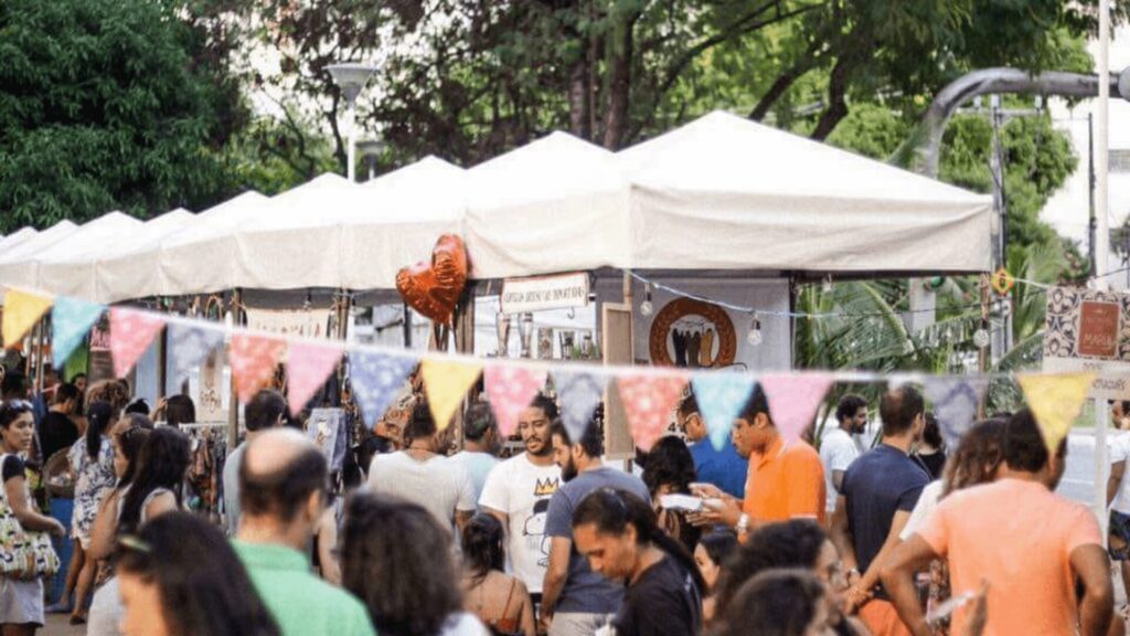 Multidão em um mercado ao ar livre com barracas e bandeiras coloridas.