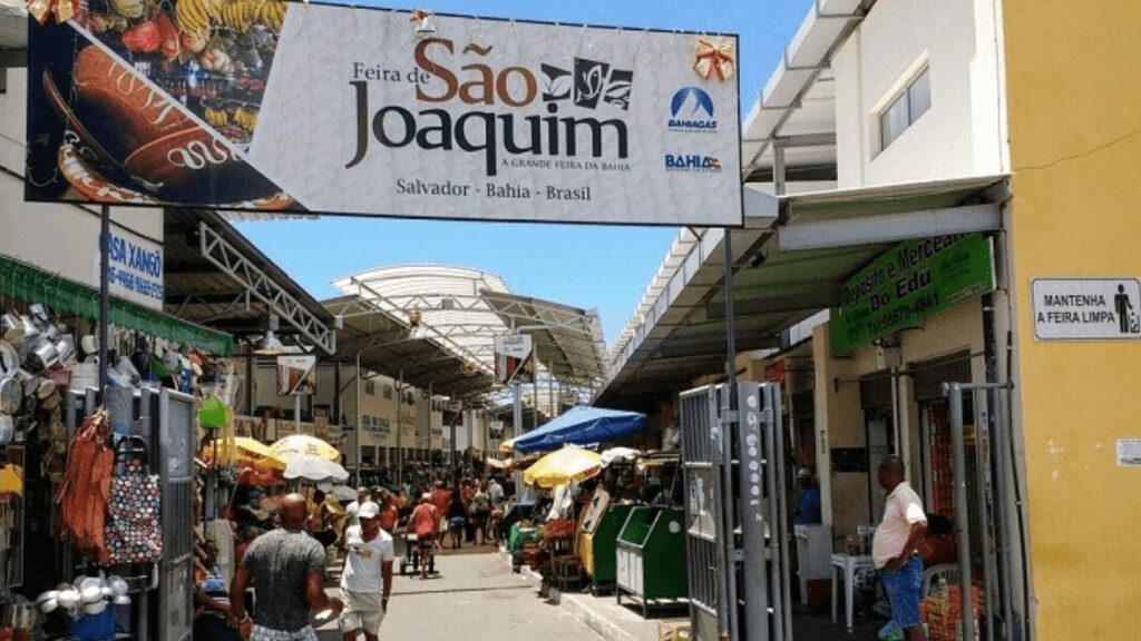 Mercado ao ar livre movimentado com pessoas caminhando, barracas com guarda-chuvas e uma placa dizendo "Feira de São Joaquim".