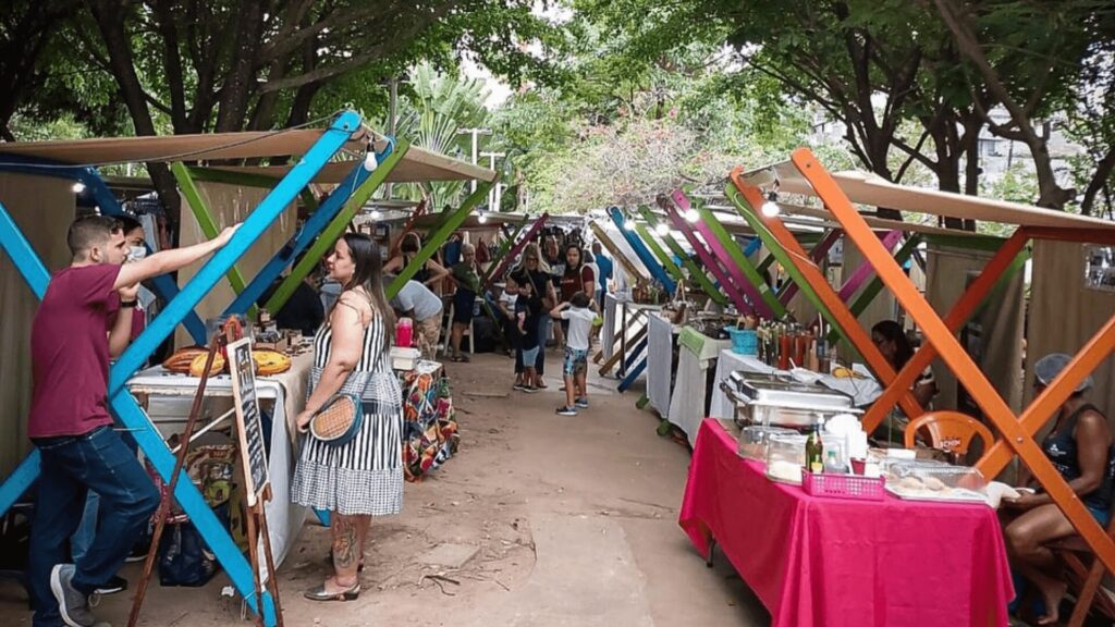 Barracas coloridas de mercado ao ar livre com artesanato e comida sob coberturas sombreadas.