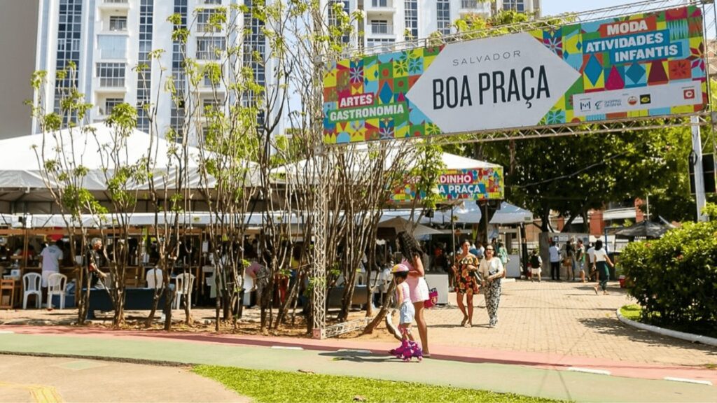 Evento de mercado ao ar livre com gente, barracas e placa da "Boa Praça" em ambiente urbano ensolarado.