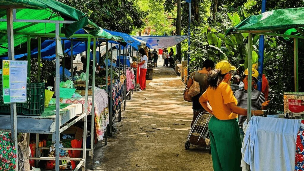 Mercado ao ar livre vibrante com barracas, lonas coloridas e vegetação exuberante ao fundo.