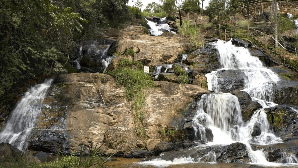 Uma série de pequenas cachoeiras desce por rochas em camadas, cercadas por vegetação tropical. Uma placa de aviso é visível no meio da cascata.