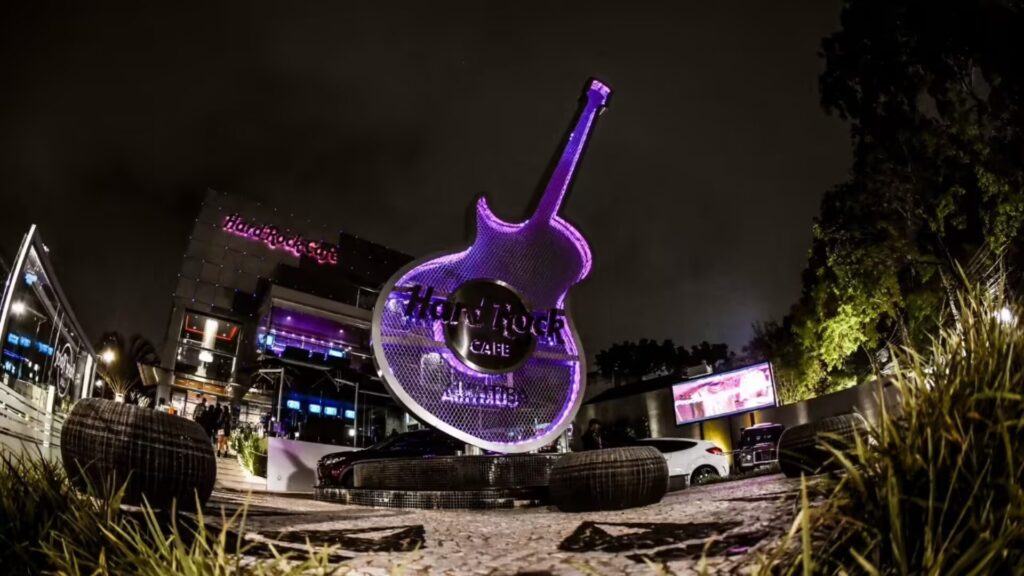 A imagem mostra o Hard Rock Cafe Curitiba à noite, com sua icônica guitarra roxa iluminada. O Hard Rock Cafe é uma rede de restaurantes temáticos com mais de 191 unidades espalhadas em 59 países, cujo tema é o Rock 'n' Roll. A atmosfera do local é um dos seus principais atrativos, com decoração inspirada na música e memorabilia de artistas famosos. O cardápio oferece opções típicas americanas, como hambúrgueres, fritas e milk-shakes. O Hard Rock Cafe Curitiba está localizado no bairro Batel, um dos mais movimentados da cidade. É um ponto turístico popular, conhecido por sua atmosfera vibrante e música ao vivo.