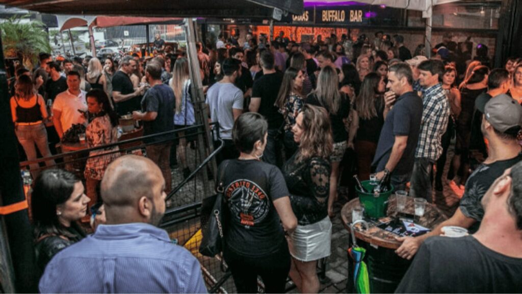 Cena de bar ao ar livre lotado com pessoas socializando e letreiros de néon.