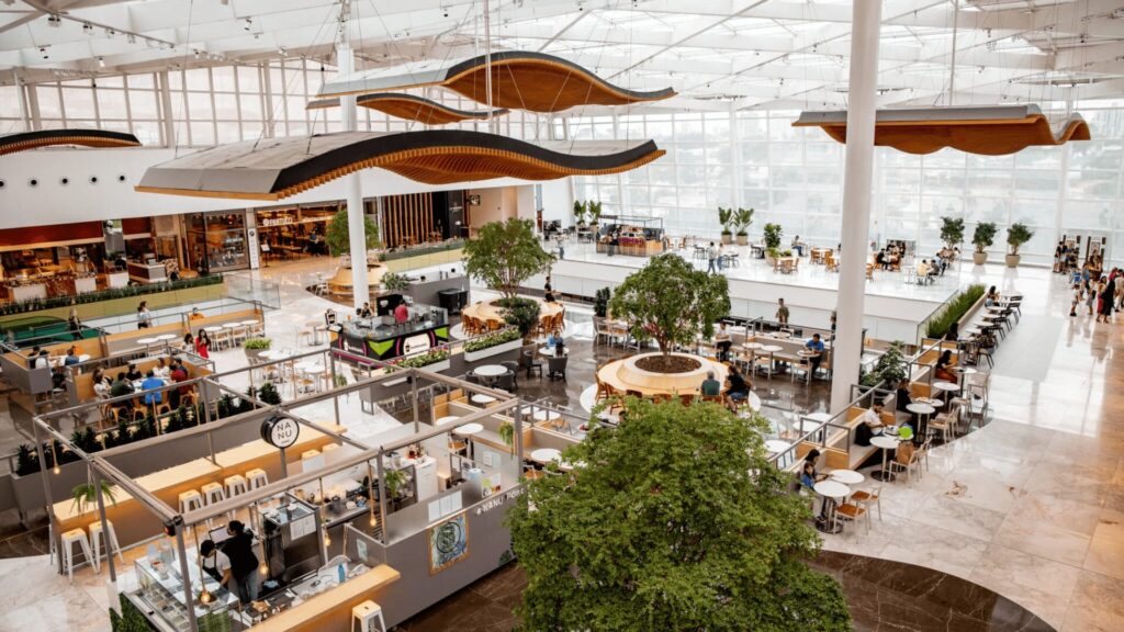 Praça de alimentação espaçosa, em vários níveis, com design moderno, áreas de estar e esculturas de madeira suspensas.