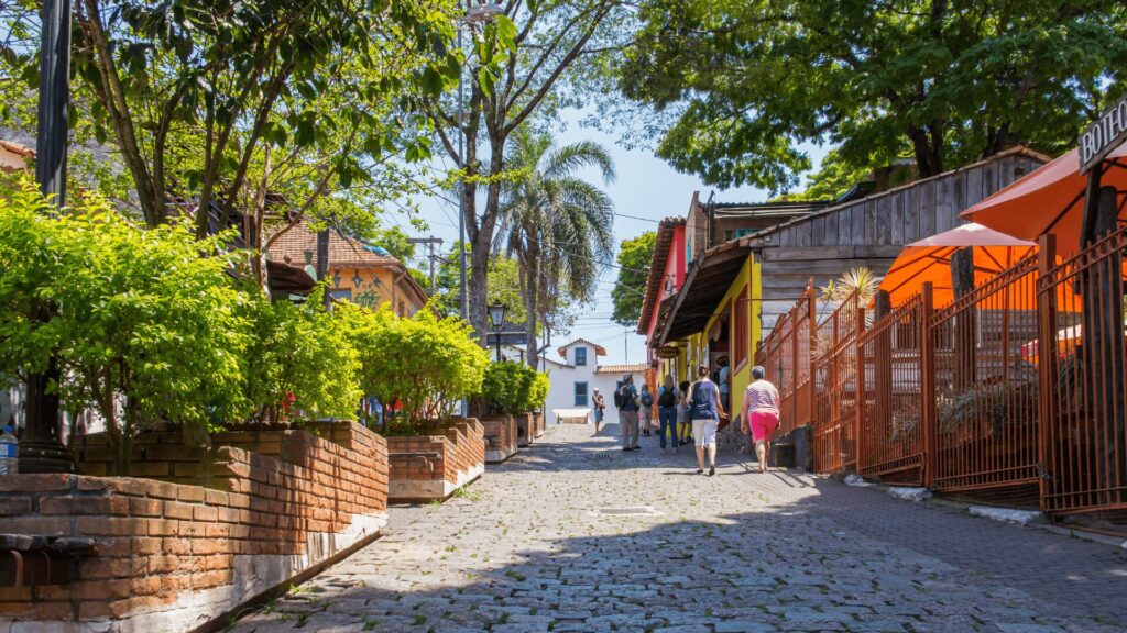 Rua de paralelepípedos com pedestres, edifícios coloridos e vegetação exuberante sob um céu claro.