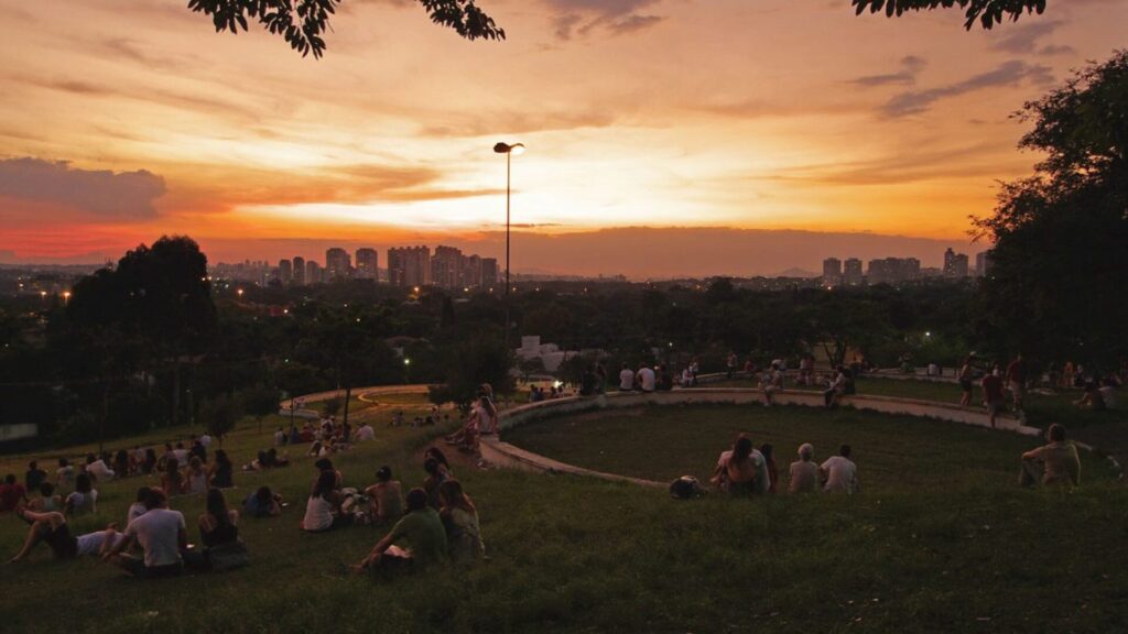 Pessoas relaxando em um parque durante o pôr do sol com a cidade ao fundo.