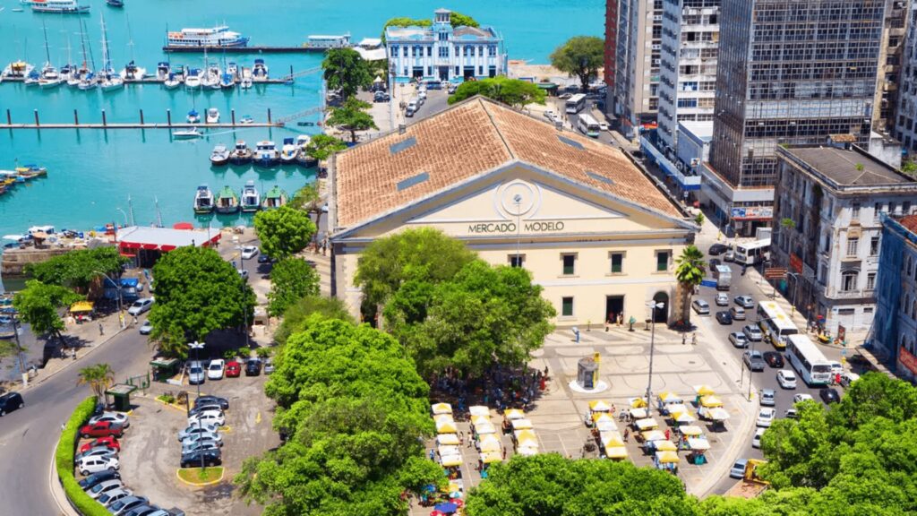 Vista aérea de uma praça de mercado movimentada perto de uma marina com barcos, cercada por edifícios e árvores.