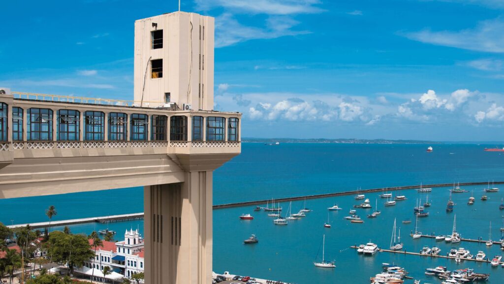 HCC (46) (1) A torre do elevador se destaca na paisagem, contrastando com os barcos menores no porto e as construções ao redor. As nuvens dispersas adicionam um elemento de dinamismo à cena, sugerindo um dia com clima variável e interessante.