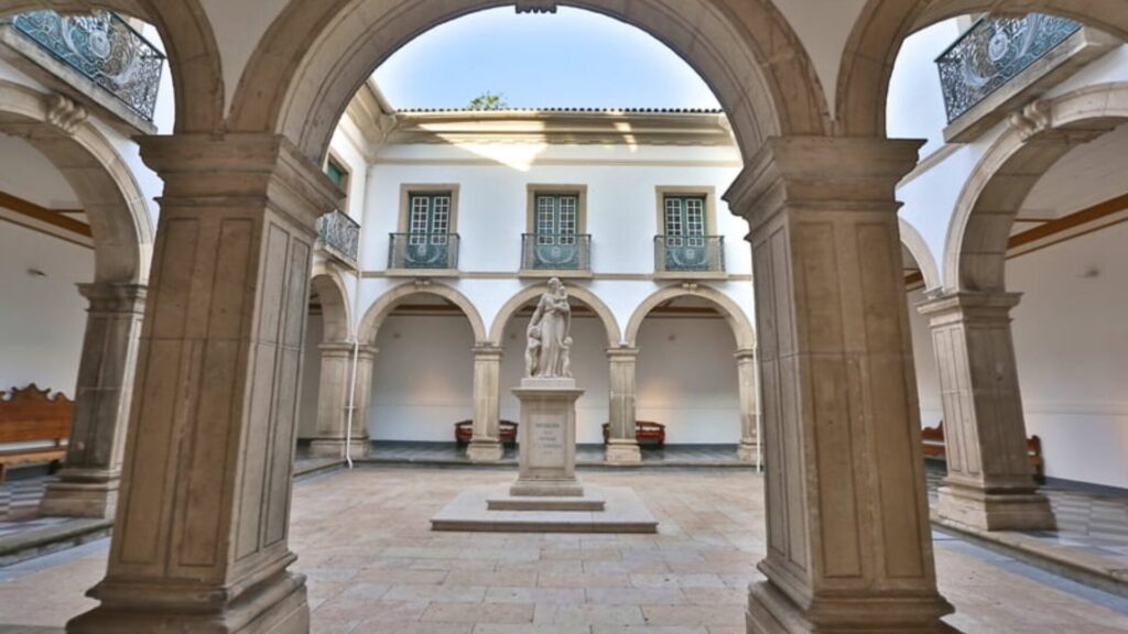 Pátio com arcos, uma estátua central e um edifício de dois andares ao fundo.