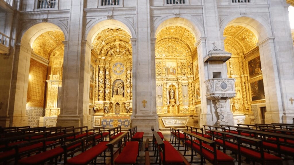 A imagem revela o interior majestoso da Catedral Basílica de Salvador, com seus altares ricamente decorados em ouro. O destaque fica para o altar principal, profusamente adornado com talhas douradas e imagens religiosas. Arcos imponentes e colunas brancas emolduram o altar, criando uma atmosfera de grandiosidade e reverência.  Fileiras de bancos de madeira com assentos vermelhos preenchem o espaço, convidando à contemplação e à oração. A iluminação suave realça os detalhes dourados e a beleza arquitetônica do local, transmitindo a riqueza histórica e artística da Catedral.