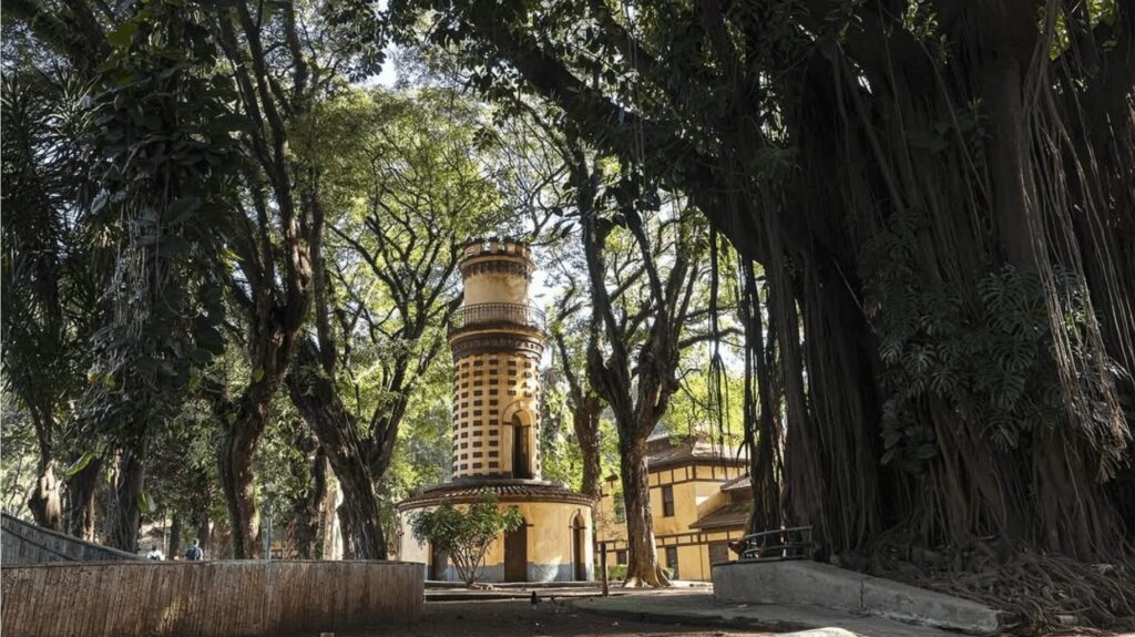 Um caminho de paralelepípedos conduz o olhar através de um bosque com árvores altas e frondosas em direção a uma torre em estilo mourisco. A torre, de cor clara com detalhes em tons terrosos, se destaca na paisagem e possui uma estrutura que lembra um minarete, com uma base circular e uma cúpula no topo. A atmosfera do local é tranquila e convidativa, um refúgio de paz em meio à natureza.