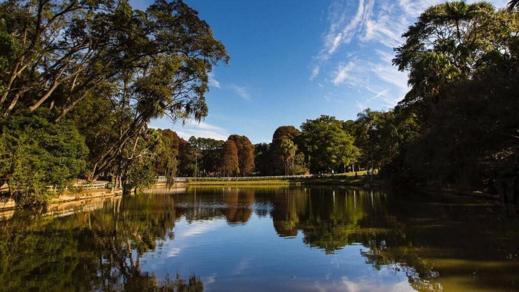 Um lago sereno reflete o azul do céu e as nuvens brancas, criando uma imagem espelhada das árvores exuberantes que o cercam. As margens do lago são adornadas por uma vegetação densa e variada, com árvores de diferentes tamanhos e tons de verde. Ao fundo, uma ponte branca e delicada convida a um passeio relaxante. A atmosfera é de paz e tranquilidade, um convite para contemplar a beleza da natureza.