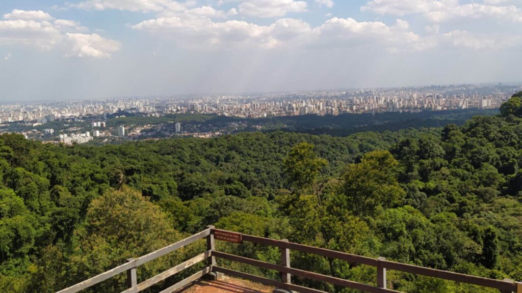 Um mirante de madeira oferece uma vista espetacular que se estende por uma vasta área urbana e uma exuberante floresta. A cidade, com seus inúmeros prédios e casas, se espalha até o horizonte, contrastando com o verde vibrante da mata que cobre as colinas. O céu azul, com algumas nuvens brancas, completa a paisagem, criando uma imagem inspiradora e que convida à contemplação.