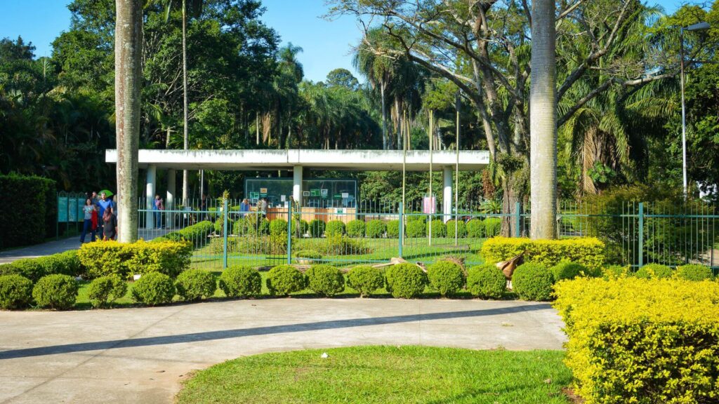 A entrada de um parque arborizado com uma guarita branca e moderna. A guarita, com um telhado plano e colunas que sustentam a estrutura, possui janelas amplas que permitem a visualização do interior.  À frente, um portão de metal com grades verdes controla o acesso ao parque.  Arbustos bem cuidados e árvores frondosas completam a cena, criando um ambiente acolhedor e convidativo.