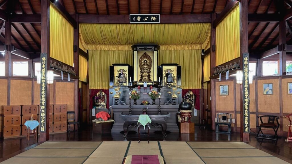 O interior de um templo budista com um altar ricamente decorado. O altar, em madeira escura, apresenta detalhes dourados e abriga estátuas de Buda e outros elementos religiosos. Cortinas amarelas e tecidos com inscrições em japonês adornam o ambiente. O chão é coberto por tatames e almofadas, criando um espaço propício à meditação e à prática religiosa. A atmosfera é de serenidade e respeito, transmitindo a paz e a harmonia características dos templos budistas.