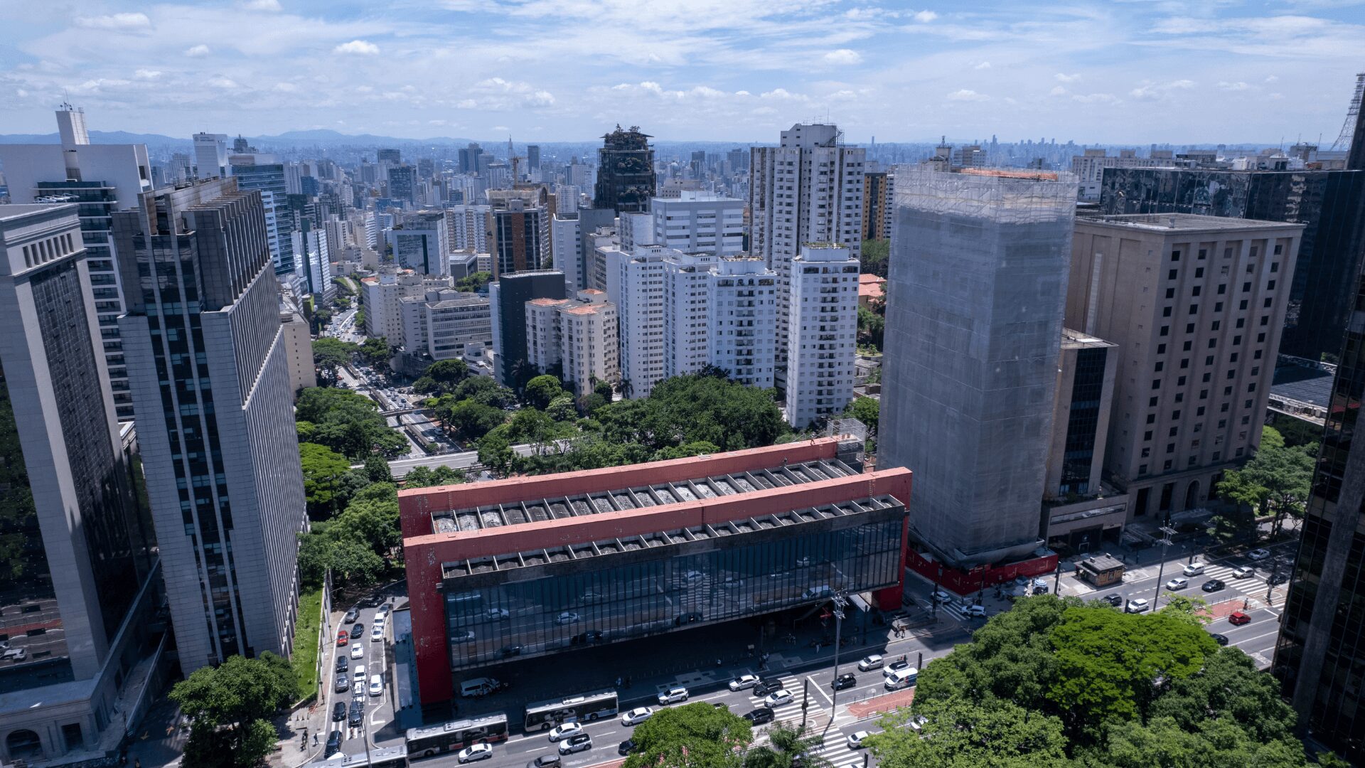 Uma vista aérea de uma metrópole vibrante, com arranha-céus imponentes e áreas verdes que se destacam em meio ao concreto. Edifícios modernos de diferentes alturas e estilos arquitetônicos dominam a paisagem, enquanto ruas movimentadas cortam a cidade em todas as direções. A presença de áreas verdes, como parques e praças, traz um toque de natureza e frescor ao ambiente urbano. Ao fundo, montanhas e um céu azul com nuvens completam a cena, criando uma imagem dinâmica e cheia de vida.