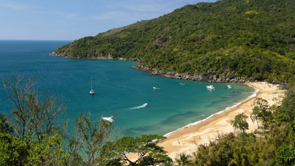 Uma praia paradisíaca de águas calmas e cristalinas, com tons de azul e verde, convidando para um mergulho. A areia é clara e fina, contrastando com o verde vibrante da vegetação que cobre as colinas ao redor.  Ao fundo, pequenas embarcações navegam tranquilamente, complementando a paisagem serena. Um lugar perfeito para relaxar e apreciar a beleza da natureza.