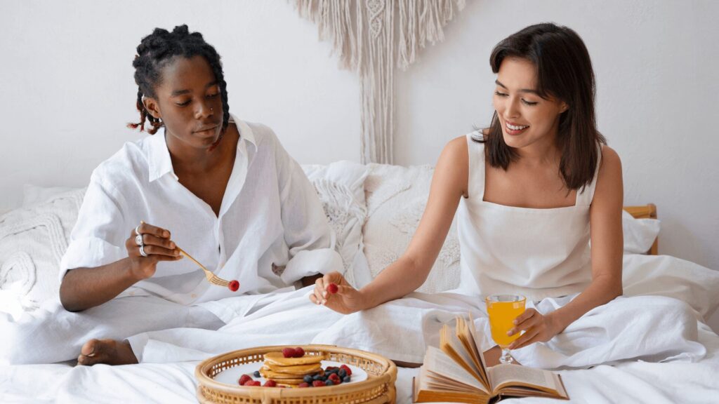 Duas pessoas em trajes brancos sentadas na cama com uma bandeja de café da manhã e um livro aberto entre elas.