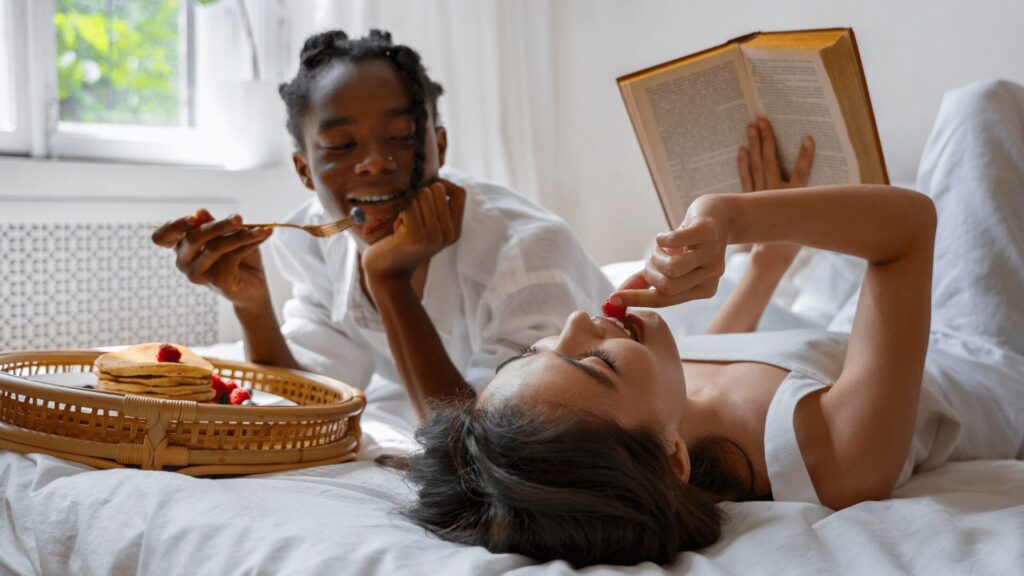 Duas pessoas na cama com café da manhã, uma delas lendo um livro.