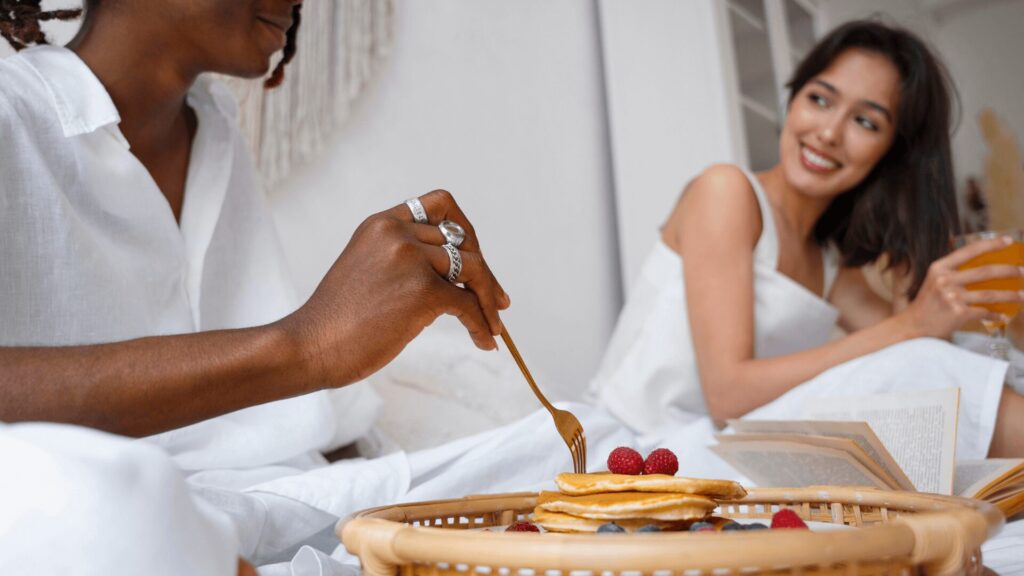 Duas pessoas aproveitando o café da manhã na cama, com panquecas e frutas.