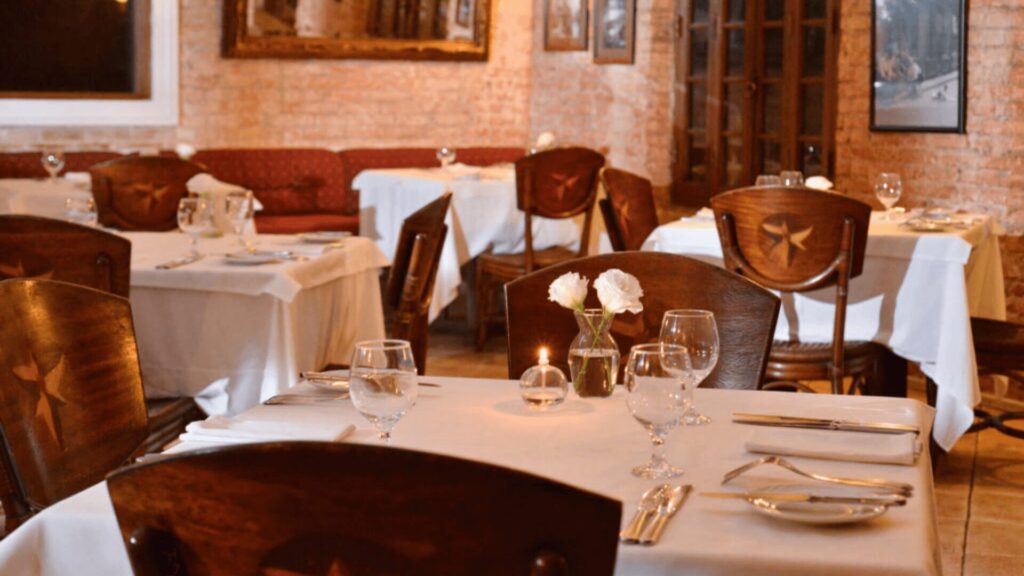 Interior aconchegante do restaurante com toalhas de mesa brancas, copos elegantes e peças centrais florais, iluminado por luz suave de velas.