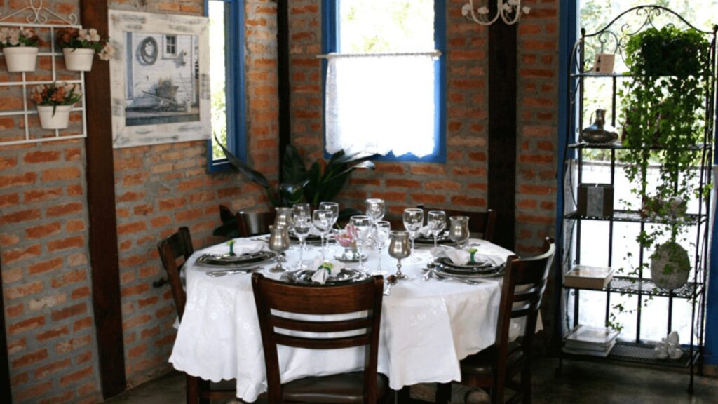 Sala de jantar aconchegante com mesa circular posta para convidados, adornada com vidros e folhagens, encostada em uma parede rústica de tijolos.