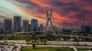 Pôr do sol sobre a icônica Ponte de São Paulo, cercada por arranha-céus modernos e uma rodovia sinuosa sob um céu colorido.