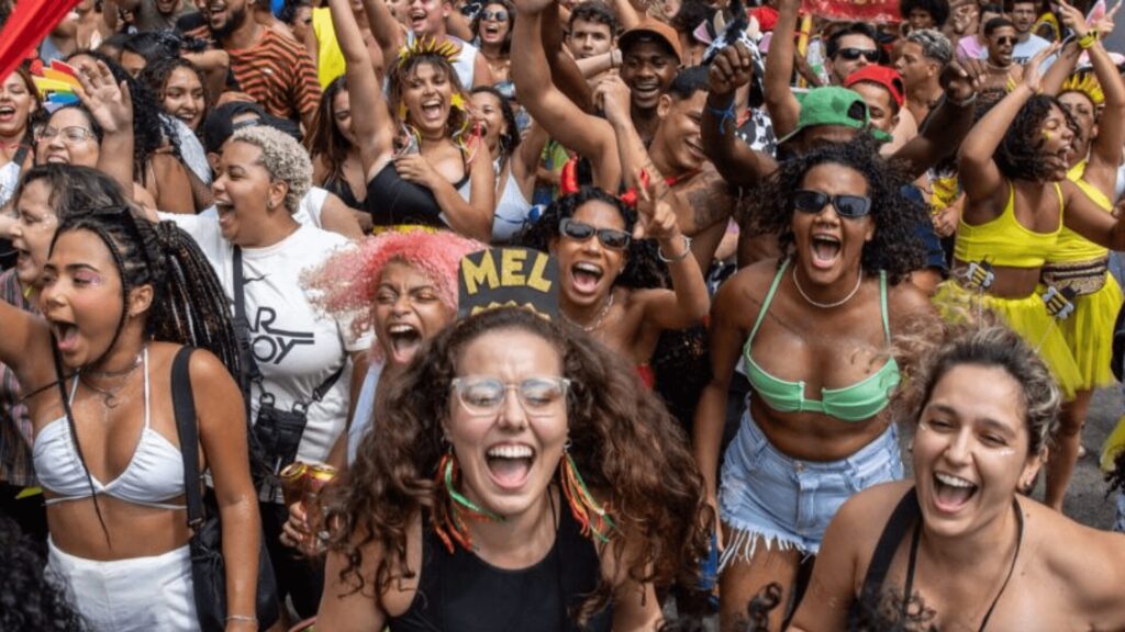 Uma multidão vibrante celebrando, vestindo roupas festivas, dançando e levantando as mãos em entusiasmo em um animado evento ao ar livre.