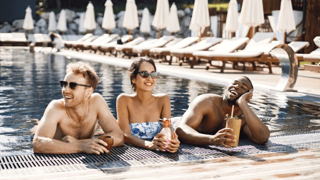 Três pessoas relaxam em uma piscina luxuosa, aproveitando drinks coloridos e rodeadas por cadeiras de lounge e guarda-sóis.