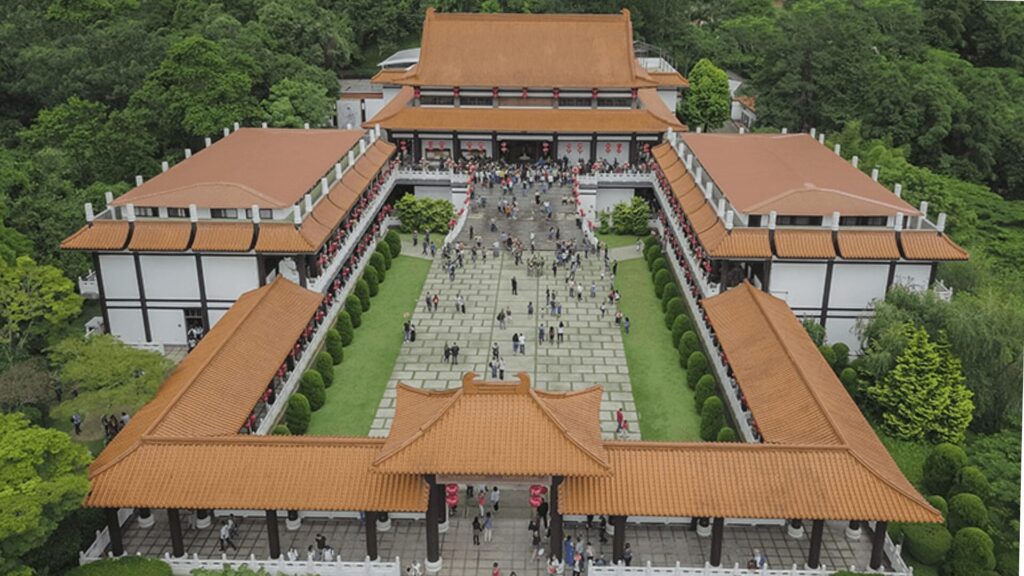 Um templo budista visto de cima, com vários prédios com telhados em camadas cor de terracota. Os prédios são brancos com detalhes dourados e estão dispostos em um padrão retangular com um pátio central. O pátio está cheio de pessoas caminhando em diferentes direções. O templo está cercado por árvores verdes.