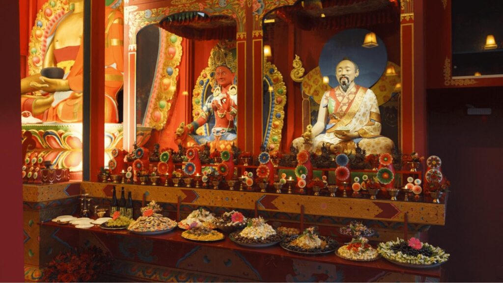 Um interior ricamente decorado de um templo budista tibetano. Duas estátuas douradas de Buda estão em altares adornados, com um Buda sentado à direita e um Buda em pé à esquerda. O Buda sentado usa um roupão branco e dourado e tem um chapéu azul atrás dele. O Buda em pé é vermelho e usa um roupão laranja e dourado. Em frente aos altares, há um longo balcão de madeira com sete pratos de oferendas. Cada prato contém uma pequena pilha de comida, como arroz, biscoitos ou frutas, e está decorado com flores e outros enfeites. O balcão é feito de madeira escura com entalhes intrincados e detalhes coloridos. As paredes e o teto do templo são pintados em cores vibrantes, com muitas decorações e detalhes. A luz no templo é suave e dourada, criando uma atmosfera pacífica e reverente.