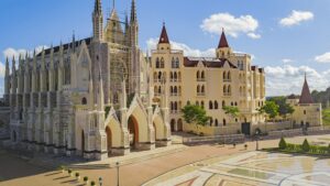 Um grande edifício de pedra em estilo gótico com torres altas e vitrais complexos. O edifício está parcialmente em construção, com andaimes visíveis. Ao lado, há um castelo de conto de fadas em tons pastéis com torres vermelhas e um pátio pavimentado com padrões geométricos. O céu está azul com nuvens brancas.