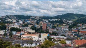 Vista panorâmica da cidade de Cotia, em São Paulo, com um céu nublado ao fundo. A cidade se estende por colinas e vales, com uma mistura de casas, edifícios e árvores. As construções variam em tamanho e estilo, com destaque para um edifício alto e marrom no centro da imagem. Uma estrada movimentada corta a cidade, com carros visíveis em movimento. As colinas ao fundo são cobertas por árvores e algumas casas. O céu é predominantemente cinza, com nuvens brancas espalhadas. A vegetação verde das árvores e dos jardins contrasta com os tons de cinza e marrom das construções.