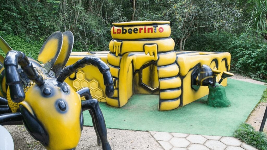 Parque infantil temático de abelhas. Uma abelha gigante de plástico preto e amarelo está em primeiro plano, com o corpo listrado e asas translúcidas. No fundo, um labirinto de blocos amarelos e pretos, com o nome "Lpbeerinto" escrito em letras brancas na parte superior. O labirinto tem buracos e túneis para as crianças brincarem. A grama verde artificial cobre o chão ao redor do labirinto, e uma trilha de pedras leva até ele. Árvores e vegetação densa cercam o parque infantil.