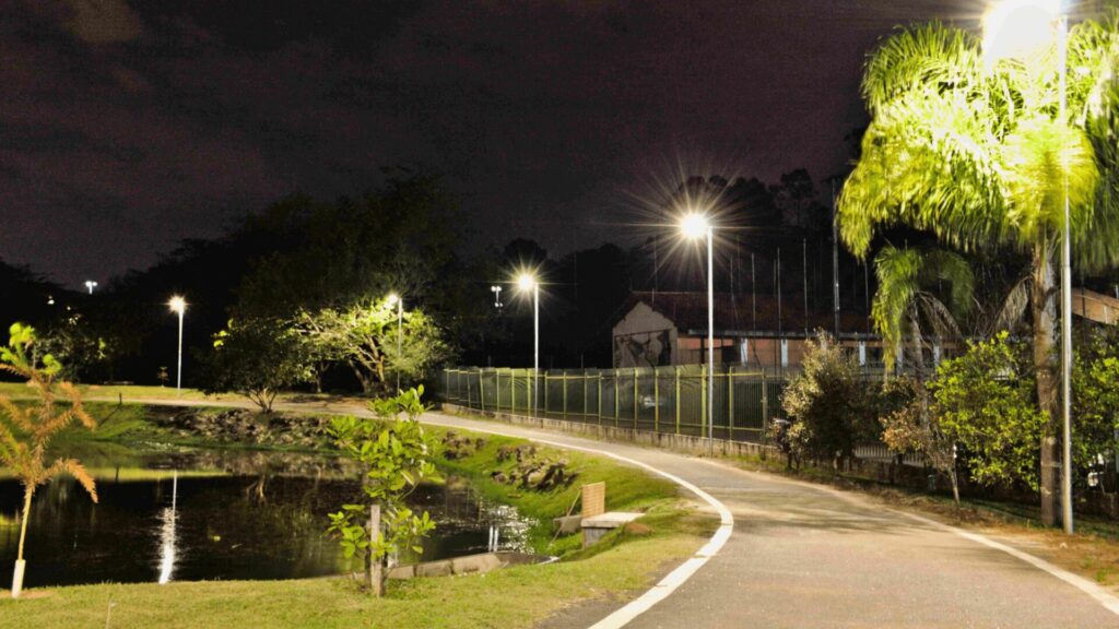 Um caminho pavimentado iluminado por postes de luz leva a um lago escuro à noite. O caminho curva-se à direita, com grama verde de cada lado. O lago reflete as luzes dos postes e as árvores ao redor. Ao fundo, um campo de esportes cercado por uma cerca de arame é visível. Palmeiras altas e outras árvores com folhagem densa se erguem à direita, sob um céu nublado. A iluminação geral é baixa, com as luzes dos postes criando pontos de luz brilhantes na cena noturna.