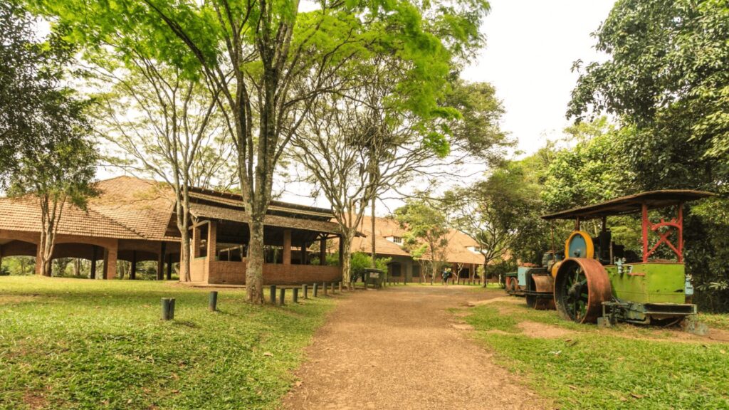 Um caminho panorâmico ladeado por árvores que passa por edifícios de tijolos e uma antiga máquina a vapor verde em um ambiente externo exuberante.